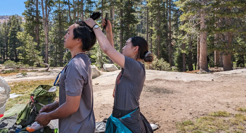 One person fixes the hair of another in a wooded area